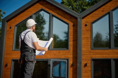 Mimar bekleme planı. Sakallı mimar elinde proje planları, yeni modern evlerin yanında duruyor ve inşaat alanında bir şeyler arıyor..