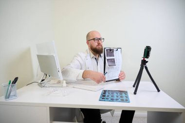 Beyaz önlüklü doktor modern klinikteki webcam üzerinden online danışmanlık yapıyor. Teletıp kavramı, uzaktan sağlık, sanal randevu, dijital sağlık.