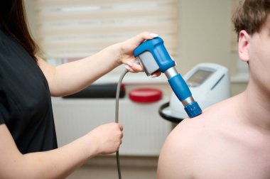 Akustik şok dalgalarıyla omuz fizyoterapisi. Doktorun arkadan şok dalgası terapisi uyguladığını yakından görmek. Kadın, ağrı kesici için pnömatik şok dalgası terapi makinesi tutuyor..