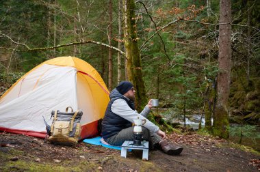 Kamp çadırının yanında oturan ve elinde sıcak kahveyle bir kupa tutan bir adam. Macera, seyahat, turizm ve kamp konsepti. Kahve içen bir adam doğada kamp yapmaktan hoşlanıyor..