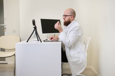 Beyaz önlüklü doktor modern klinikteki webcam üzerinden online danışmanlık yapıyor. Teletıp kavramı, uzaktan sağlık, sanal randevu, dijital sağlık.