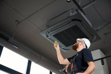 Sakallı adam merdivende duruyor ofise klima yerleştiriyor. Modern binada tavana monte edilmiş klima. Çatı katı tarzı. Endüstriyel iç tasarım. Teknisyen servisi.