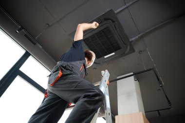 Sakallı adam merdivende duruyor ofise klima yerleştiriyor. Modern binada tavana monte edilmiş klima. Çatı katı tarzı. Endüstriyel iç tasarım. Teknisyen servisi.