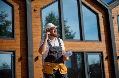 Mimari bekleme planı ve akıllı telefondan konuşma. Sakallı mimar elinde proje tasarısı, kağıt planları ile inşaat sahasına bakan yeni modern evlerin kombinasyonunun yanında duruyor..