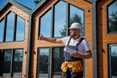 Mimar bekleme planı. Sakallı mimar elinde proje planları, yeni modern evlerin yanında duruyor ve inşaat alanında bir şeyler arıyor..