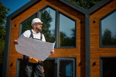 Mimar bekleme planı. Sakallı mimar elinde proje planları, yeni modern evlerin yanında duruyor ve inşaat alanında bir şeyler arıyor..