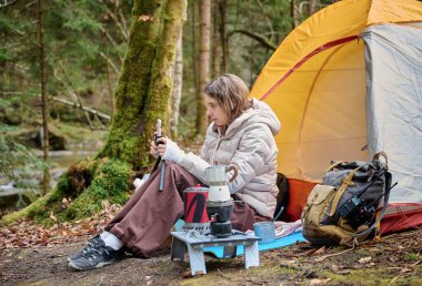Güzel bir blogcu kız ormanda çadırın yanında oturur ve akıllı telefon kamerasıyla kendini filme çeker. Gezgin blogcu kadın kamp çadırının yakınında sosyal video hikayesi yazıyor. açık hava vlog kaydedicisi