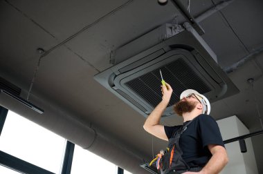 Sakallı adam merdivende duruyor ofise klima yerleştiriyor. Modern binada tavana monte edilmiş klima. Çatı katı tarzı. Endüstriyel iç tasarım. Teknisyen servisi.