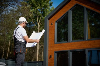 Mimar bekleme planı. Sakallı mimar elinde proje planları, yeni modern evlerin yanında duruyor ve inşaat alanında bir şeyler arıyor..