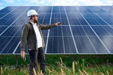 Güneş istasyonu yakınında koruyucu kasklı üniformalı bir mühendis telsiz tutuyor. Denetmen güneş panellerini güneş çiftliğindeki radyo istasyonuyla kontrol ediyor. Güneş ve yenilenebilir enerji kavramı.