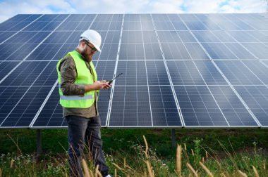 Güneş istasyonu yakınında koruyucu kasklı üniformalı bir mühendis akıllı telefon ve telsiz tutuyor. Denetmen güneş panellerini güneş çiftliğindeki akıllı telefonuyla kontrol ediyor. Güneş ve yenilenebilir enerji