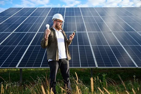 Üniformalı mühendis. Güneş istasyonu yakınlarında koruyucu kasklı. Elinde ampul var. Denetmen güneş panellerini güneş çiftliğindeki akıllı telefonuyla kontrol ediyor. Güneş ve yenilenebilir enerji kavramı.
