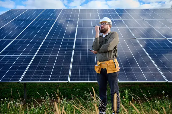 Güneş istasyonu yakınında koruyucu kasklı üniformalı bir mühendis telsiz tutuyor. Denetmen güneş panellerini güneş çiftliğindeki radyo istasyonuyla kontrol ediyor. Güneş ve yenilenebilir enerji kavramı.