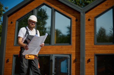 Mimar bekleme planı. Sakallı mimar elinde proje planları, yeni modern evlerin yanında duruyor ve inşaat alanında bir şeyler arıyor..