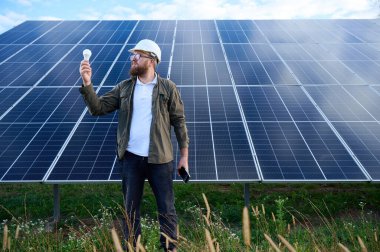 Üniformalı mühendis. Güneş istasyonu yakınlarında koruyucu kasklı. Elinde ampul var. Denetmen güneş panellerini güneş çiftliğindeki akıllı telefonuyla kontrol ediyor. Güneş ve yenilenebilir enerji kavramı.