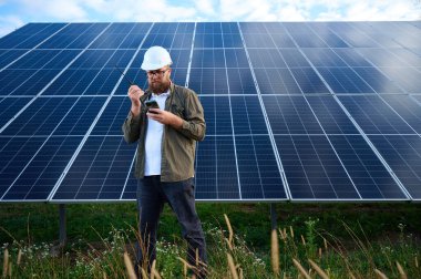 Güneş istasyonu yakınında koruyucu kasklı üniformalı bir mühendis akıllı telefon ve telsiz tutuyor. Denetmen güneş panellerini güneş çiftliğindeki akıllı telefonuyla kontrol ediyor. Güneş ve yenilenebilir enerji