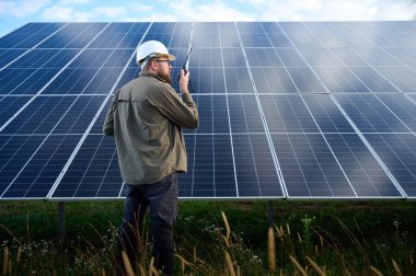 Güneş istasyonu yakınında koruyucu kasklı üniformalı bir mühendis telsiz tutuyor. Denetmen güneş panellerini güneş çiftliğindeki radyo istasyonuyla kontrol ediyor. Güneş ve yenilenebilir enerji kavramı.