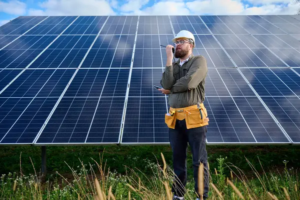 Güneş istasyonu yakınında koruyucu kasklı üniformalı bir mühendis telsiz tutuyor. Denetmen güneş panellerini güneş çiftliğindeki radyo istasyonuyla kontrol ediyor. Güneş ve yenilenebilir enerji kavramı.