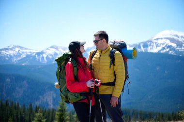 İki yürüyüşçü bir dağ sırtında mola veriyor. Sırt çantalı sportif çift, karlı dağların arka planında duruyorlar..