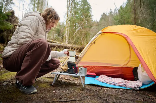 Yürüyüşçü kadın kamp çadırının yanında kahve hazırlıyor. İlkbaharın başlarında ormanda bir bayan gezgin sıcak kahveyi bir turist bardağında tutuyor..