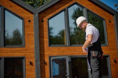 Mimar bekleme planı. Sakallı mimar elinde proje planları, yeni modern evlerin yanında duruyor ve inşaat alanında bir şeyler arıyor..