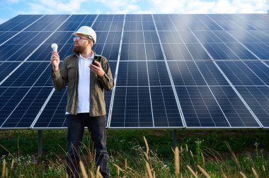 Üniformalı mühendis. Güneş istasyonu yakınlarında koruyucu kasklı. Elinde ampul var. Denetmen güneş panellerini güneş çiftliğindeki akıllı telefonuyla kontrol ediyor. Güneş ve yenilenebilir enerji kavramı.