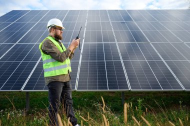 Güneş istasyonu yakınında koruyucu kasklı üniformalı bir mühendis akıllı telefon ve telsiz tutuyor. Denetmen güneş panellerini güneş çiftliğindeki akıllı telefonuyla kontrol ediyor. Güneş ve yenilenebilir enerji