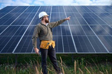 Güneş istasyonu yakınında koruyucu kasklı üniformalı bir mühendis telsiz tutuyor. Denetmen güneş panellerini güneş çiftliğindeki radyo istasyonuyla kontrol ediyor. Güneş ve yenilenebilir enerji kavramı.