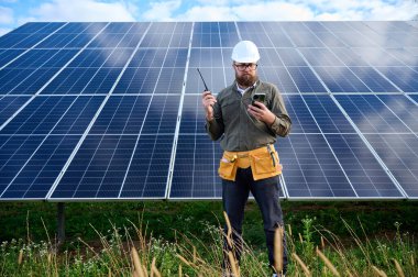 Güneş istasyonu yakınında koruyucu kasklı üniformalı bir mühendis akıllı telefon ve telsiz tutuyor. Denetmen güneş panellerini güneş çiftliğindeki akıllı telefonuyla kontrol ediyor. Güneş ve yenilenebilir enerji