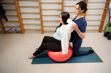 Hamile kadın, fitness topu kullanarak fizyoterapistle doğum öncesi egzersizleri yapıyor. Hamile müşteriye hamilelik fizyoterapi seansında rehberlik eden kadın uzman..