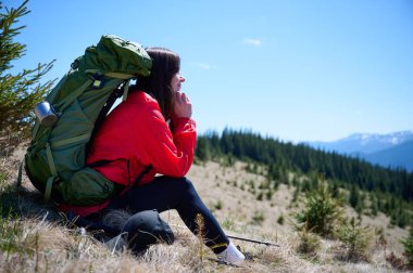 Çimenli dağ yamacında yürüyüş direğiyle dinlenen kadın yürüyüşçü. Kırmızı ceketli kadın, karlı tepelerin ve güneşli havanın tadını çıkarıyor. Açık hava seyahati ve macera yaşam tarzı.