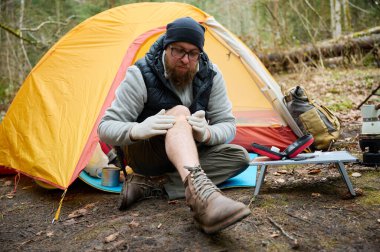 Ormandaki çadırın yakınındaki ilk yardım çantasını kullanan yaralı bacaklı bir yürüyüşçü. Stresli bir yürüyüşçü dağda kırık diziyle tek başına şikayet ediyor..