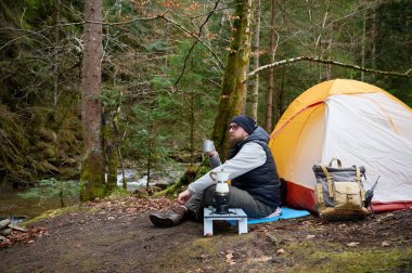Kamp çadırının yanında oturan ve elinde sıcak kahveyle bir kupa tutan bir adam. Macera, seyahat, turizm ve kamp konsepti. Kahve içen bir adam doğada kamp yapmaktan hoşlanıyor..