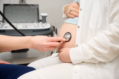 Tıbbi muayene sırasında hamile bir kadının karnını steteskopla dinleyen doktorun görüntüsü. Kadın doktor, hamile kadının karnını steteskopla kontrol ediyor. Hamilelik sırasında tıbbi bakım