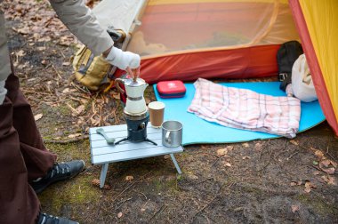 Yürüyüşçü kadın kamp çadırının yanında kahve hazırlıyor. İlkbaharın başlarında ormanda bir bayan gezgin sıcak kahveyi bir turist bardağında tutuyor..
