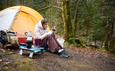 Güzel bir blogcu kız ormanda çadırın yanında oturur ve akıllı telefon kamerasıyla kendini filme çeker. Gezgin blogcu kadın kamp çadırının yakınında sosyal video hikayesi yazıyor. açık hava vlog kaydedicisi