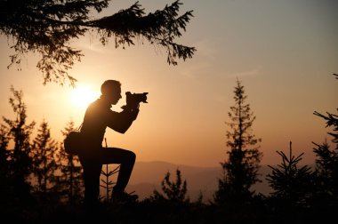 Gün batımında dağlarda elinde kamera olan bir adamın silueti. Doğadaki fotoğrafçı, batan güneşin sıcak altın ışığıyla arka planda..