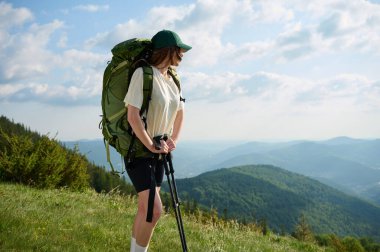 Kadın yürüyüşçü çimenli bir tepede duruyor, engin dağ manzarasını seyrediyor. Şapka ve şort giyen maceraperest, büyük bir sırt çantası taşıyor ve yürüyüş direkleri kullanıyor..