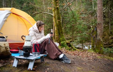 Güzel bir blogcu kız ormanda çadırın yanında oturur ve akıllı telefon kamerasıyla kendini filme çeker. Gezgin blogcu kadın kamp çadırının yakınında sosyal video hikayesi yazıyor. açık hava vlog kaydedicisi