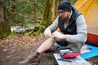 Ormandaki çadırın yakınındaki ilk yardım çantasını kullanan yaralı bacaklı bir yürüyüşçü. Stresli bir yürüyüşçü dağda kırık diziyle tek başına şikayet ediyor..