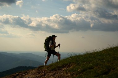 Yürüyüşçü gün batımında çimli dağ tepelerine tırmanır. Kadın altın ışıkta yokuş yukarı yürüyor, arkasında bulutlar var..