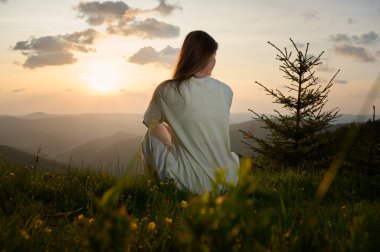 Kadın huzur içinde çimenli bir tepede oturur altın ışığın manzarayı yıkamasını seyreder. Yuvarlanan dağlar ve yemyeşil bir ağaç akşamları sakin bir atmosfer yaratır..