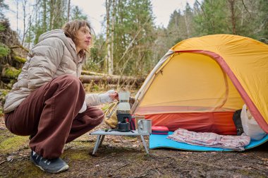 Yürüyüşçü kadın kamp çadırının yanında kahve hazırlıyor. İlkbaharın başlarında ormanda bir bayan gezgin sıcak kahveyi bir turist bardağında tutuyor..
