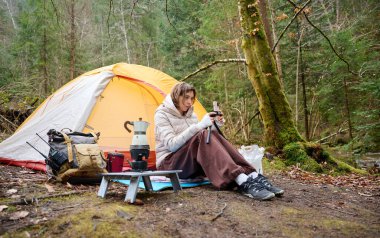 Güzel bir blogcu kız ormanda çadırın yanında oturur ve akıllı telefon kamerasıyla kendini filme çeker. Gezgin blogcu kadın kamp çadırının yakınında sosyal video hikayesi yazıyor. açık hava vlog kaydedicisi
