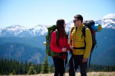 İki yürüyüşçü bir dağ sırtında mola veriyor. Sırt çantalı sportif çift, karlı dağların arka planında duruyorlar..
