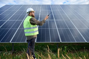 Güneş istasyonu yakınında koruyucu kasklı üniformalı bir mühendis telsiz tutuyor. Denetmen güneş panellerini güneş çiftliğindeki radyo istasyonuyla kontrol ediyor. Güneş ve yenilenebilir enerji kavramı.
