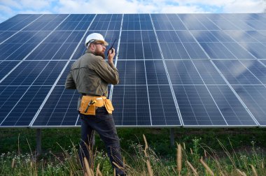 Güneş istasyonu yakınında koruyucu kasklı üniformalı bir mühendis telsiz tutuyor. Denetmen güneş panellerini güneş çiftliğindeki radyo istasyonuyla kontrol ediyor. Güneş ve yenilenebilir enerji kavramı.