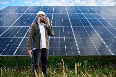 Güneş istasyonu yakınında koruyucu kasklı üniformalı bir mühendis telsiz tutuyor. Denetmen güneş panellerini güneş çiftliğindeki radyo istasyonuyla kontrol ediyor. Güneş ve yenilenebilir enerji kavramı.