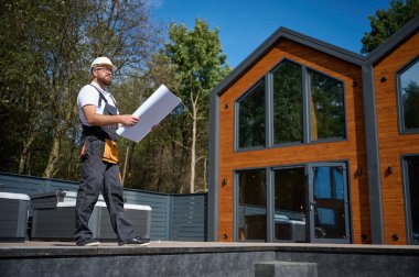 Mimar bekleme planı. Sakallı mimar elinde proje planları, yeni modern evlerin yanında duruyor ve inşaat alanında bir şeyler arıyor..