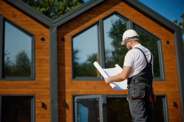 Mimar bekleme planı. Sakallı mimar elinde proje planları, yeni modern evlerin yanında duruyor ve inşaat alanında bir şeyler arıyor..